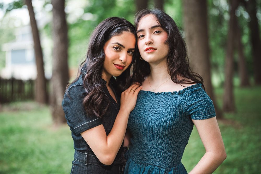 Two people in dark dresses pose together outdoors in a wooded setting with soft natural lighting.