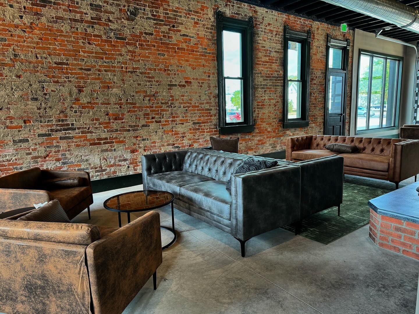 Rustic industrial loft interior with exposed brick walls, tufted leather sofa, and vintage wooden chairs in natural light.