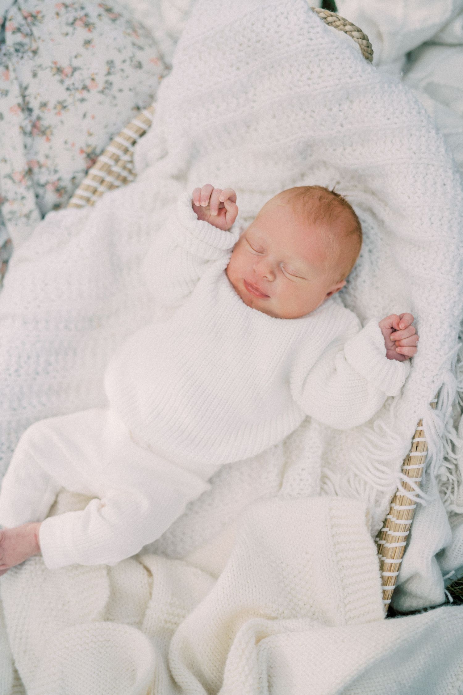 A sleeping baby wearing a soft white knit outfit rests on white blankets against floral bedding.