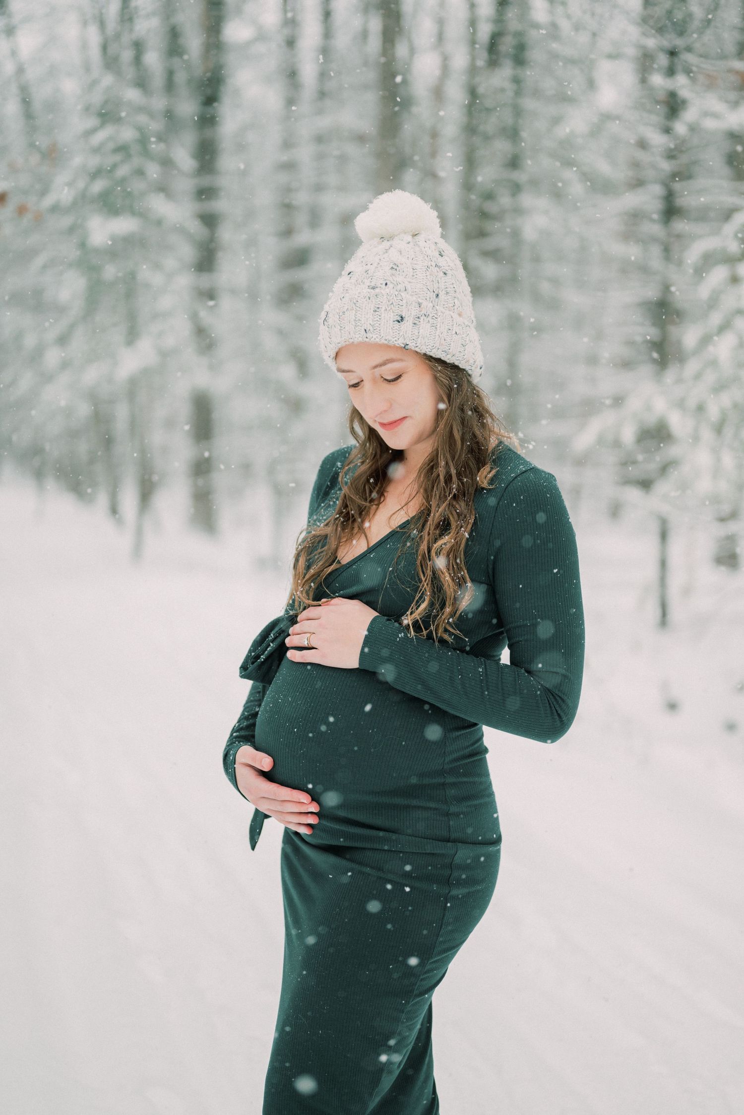A serene winter maternity portrait with falling snow and forest backdrop.