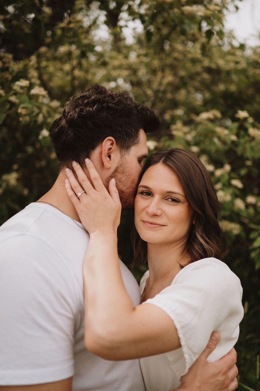 A tender embrace between a couple in white clothing against a backdrop of lush greenery.