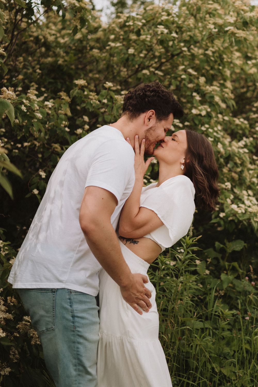 A romantic kiss shared between a couple in white clothing surrounded by blooming spring foliage.