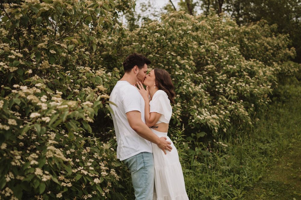 A couple shares a passionate kiss along a nature trail with flowering bushes.