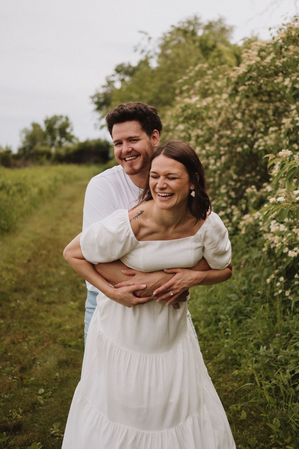 A joyful couple laughs while embracing on a grassy hillside with blooming elderflower bushes.