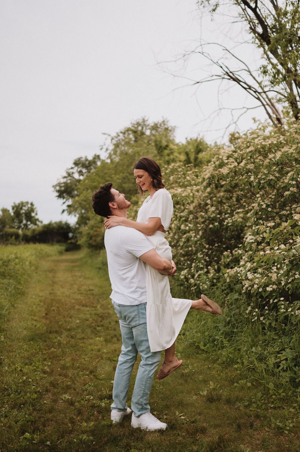 A playful lift between a couple on a countryside path lined with flowering bushes.