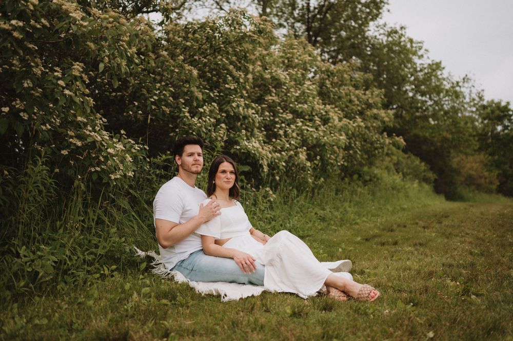 A romantic outdoor photoshoot of a couple sitting on a blanket in a grassy field with blooming trees in the background.