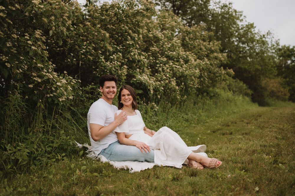 A couple shares an intimate moment while sitting on a white blanket in a sun-dappled meadow.