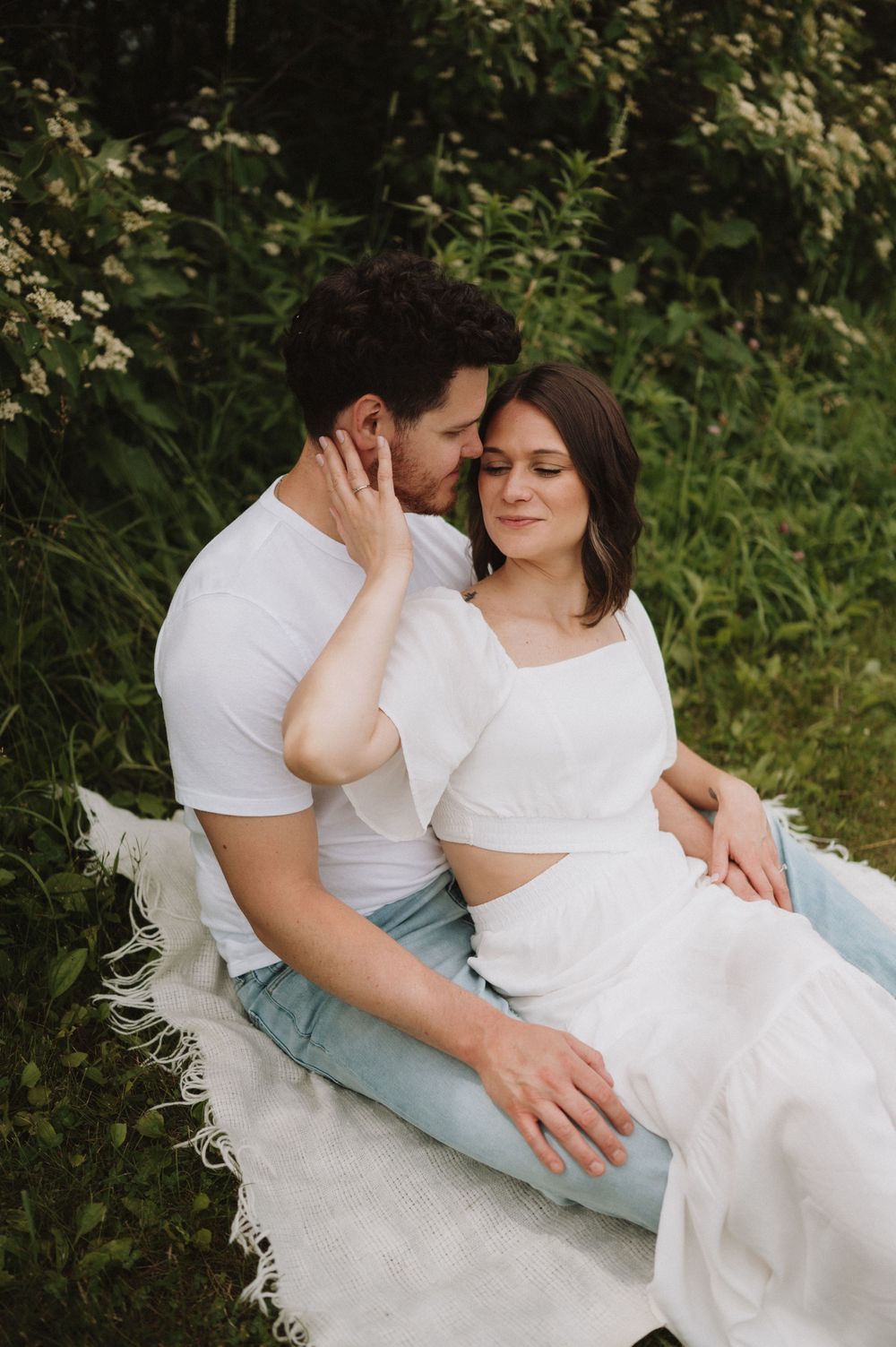 A romantic outdoor portrait of a couple in white clothing sitting on a blanket surrounded by greenery.