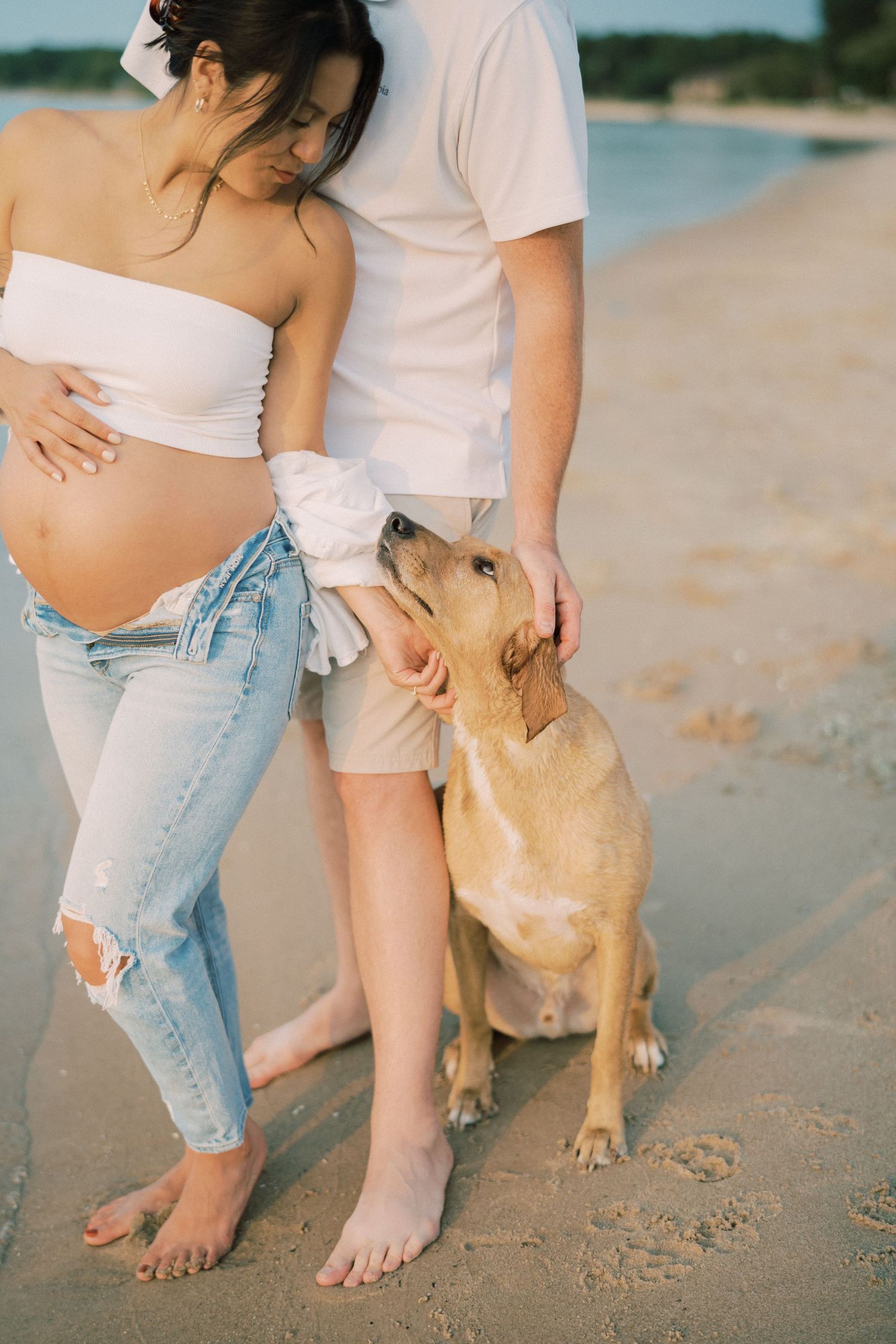 A couple in casual white attire stands on a beach with their yellow Labrador retriever, enjoying a sunny day.