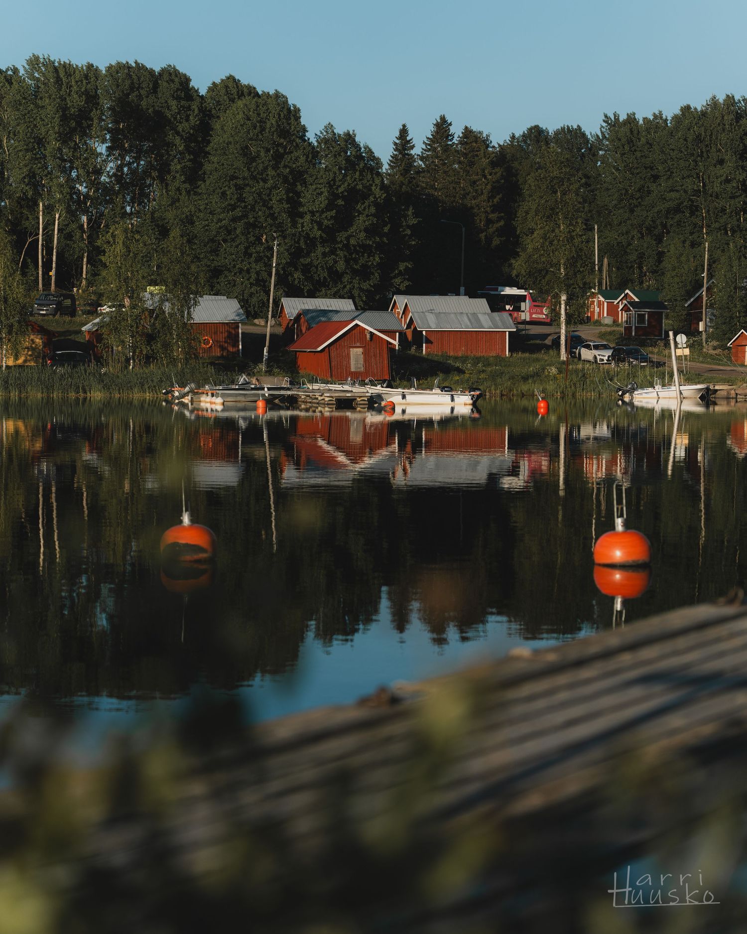 Red cabins and boats reflect perfectly in a calm lake with orange buoys floating on the surface.