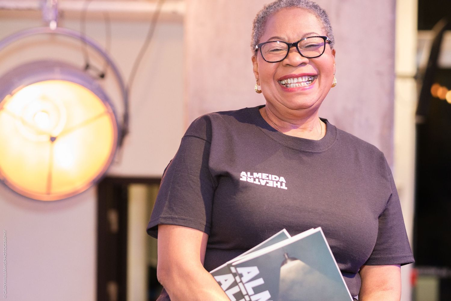 Staff member in black t-shirt holding books in a warmly lit restaurant setting.
