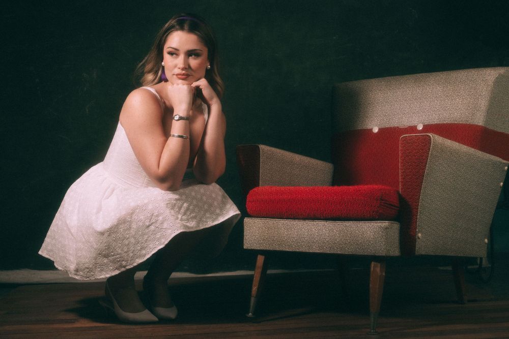 Artistic portrait in white dress next to mid-century modern chair.
