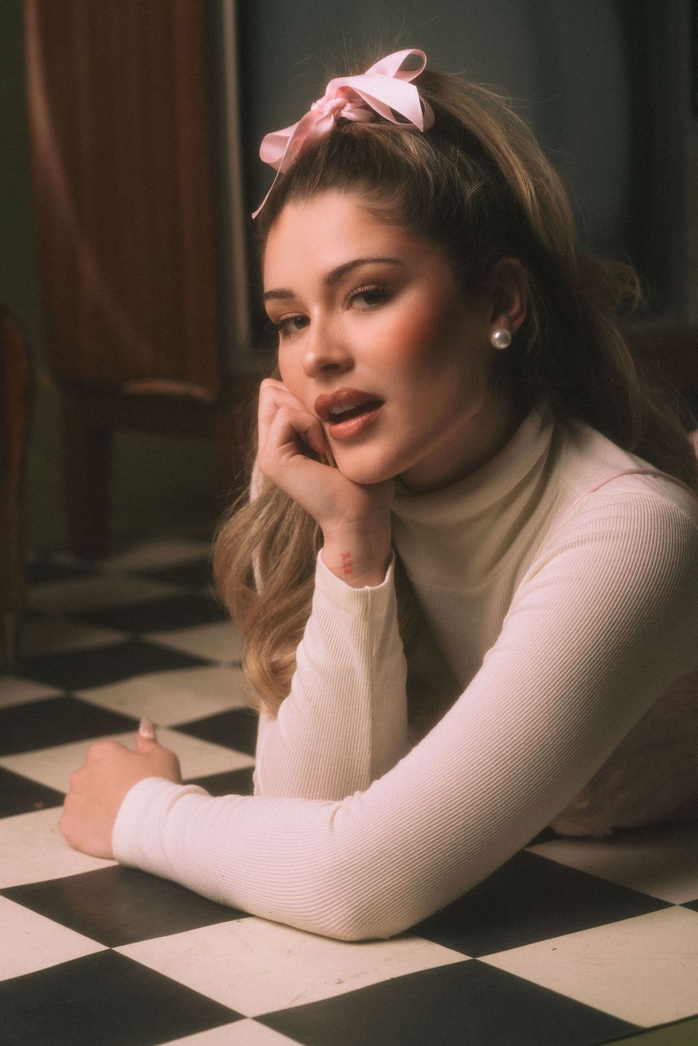 Retro-styled fashion portrait with pink hair bow and cream turtleneck sweater on checkered floor.