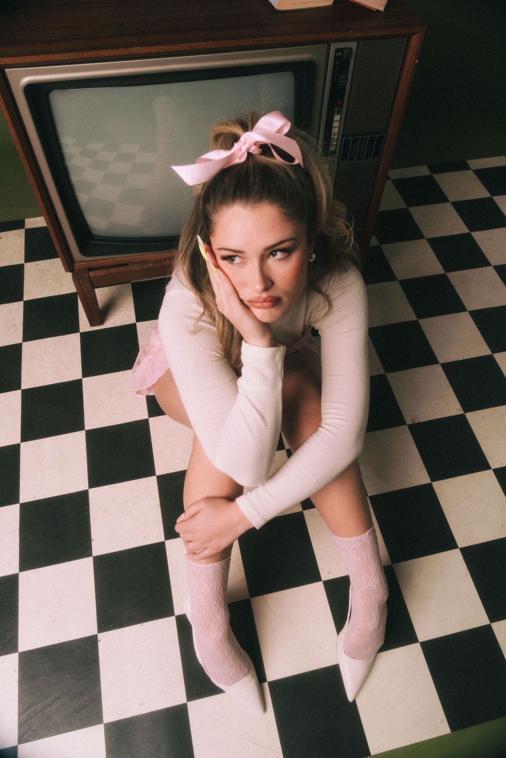 Woman in white sweater and pink bow lying on checkered floor near vintage TV.