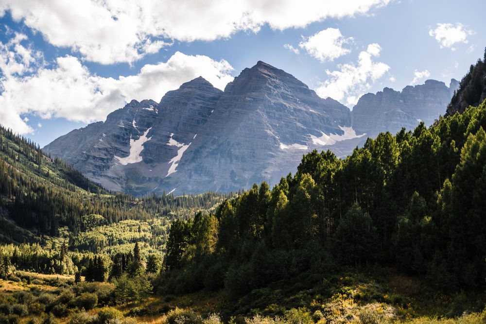Majestic mountain peaks rise above a forested valley under blue skies with white clouds.