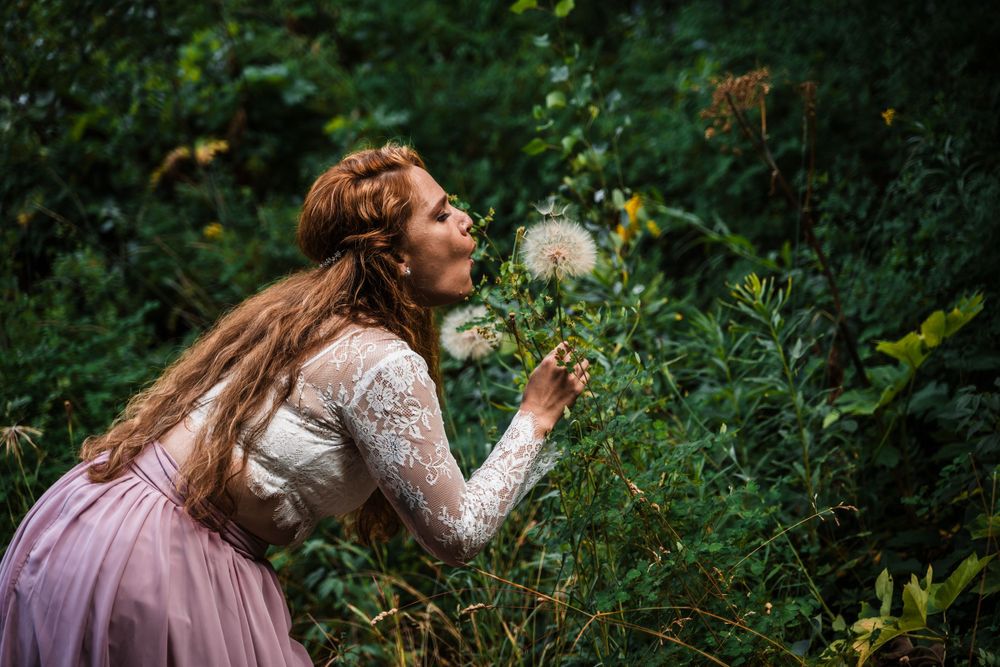 Ethereal nature scene of someone blowing on a dandelion in a lace dress among green foliage.