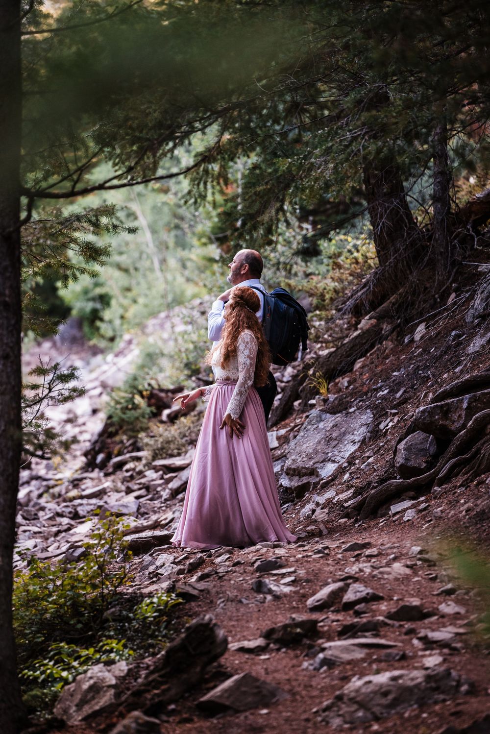 Figure in flowing pink dress walks rocky mountain trail through forest setting.