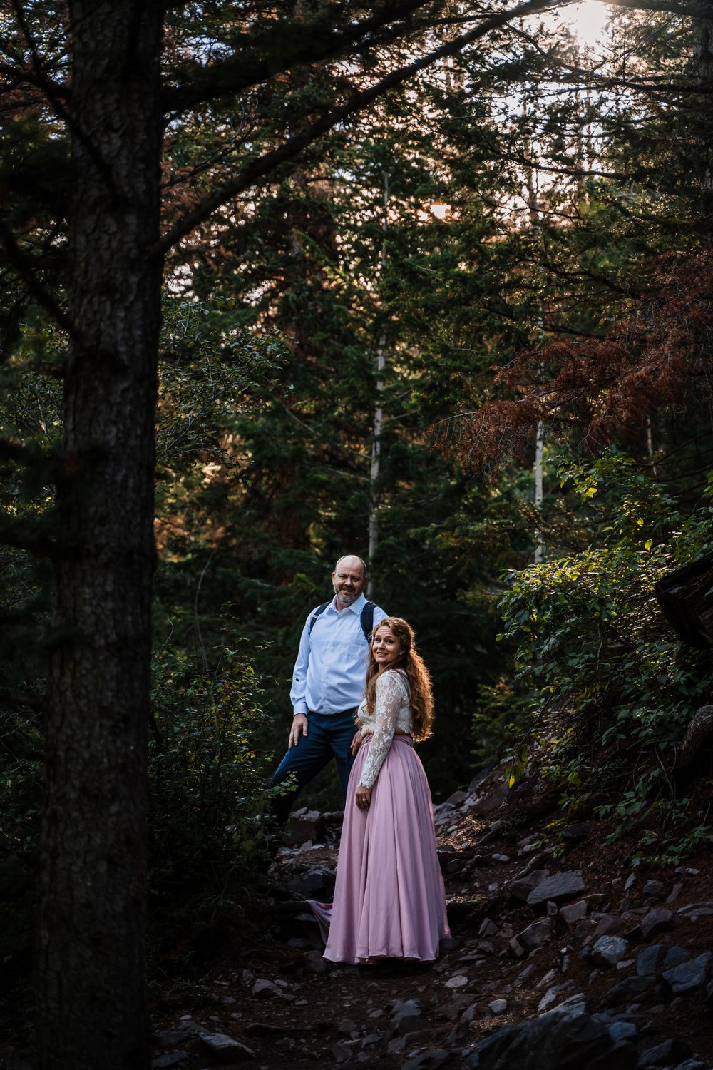 Couple poses on forested mountain trail during sunset photoshoot.