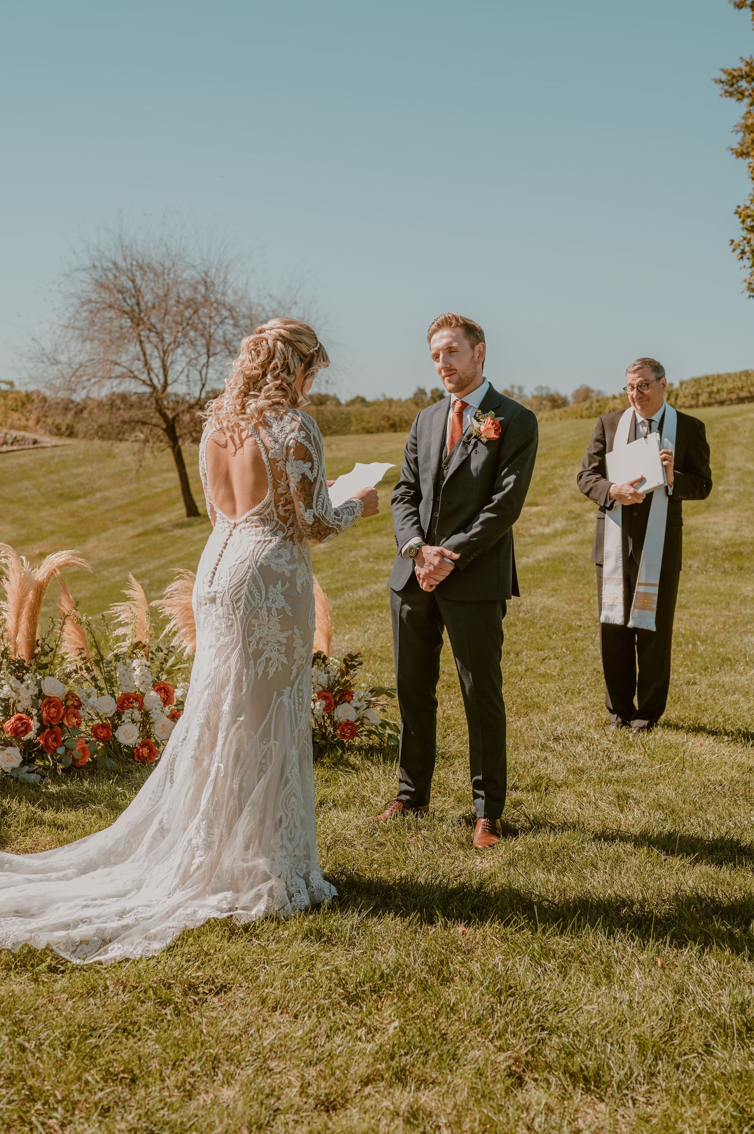 An outdoor wedding ceremony on a grassy hill with a trailing lace dress, grey suit and officiant against blue skies. By Samantha Campbell.