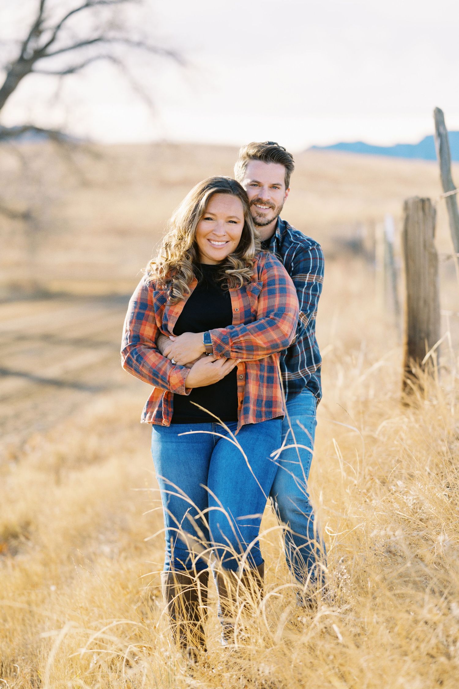 Couple posing together in rustic field with mountains in background.
