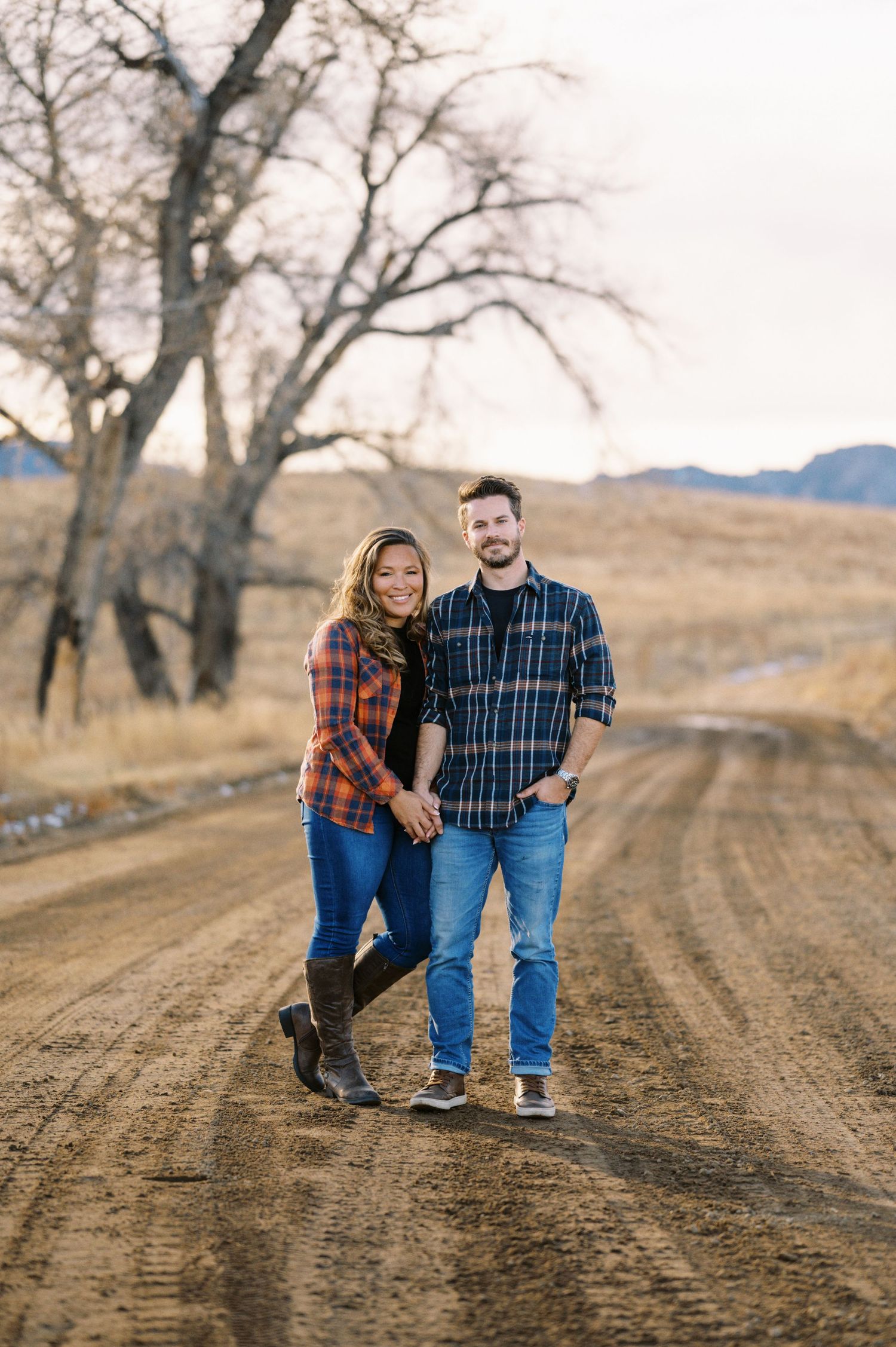 A couple in matching plaid shirts stands together on a dirt road with mountains and bare trees in the background.