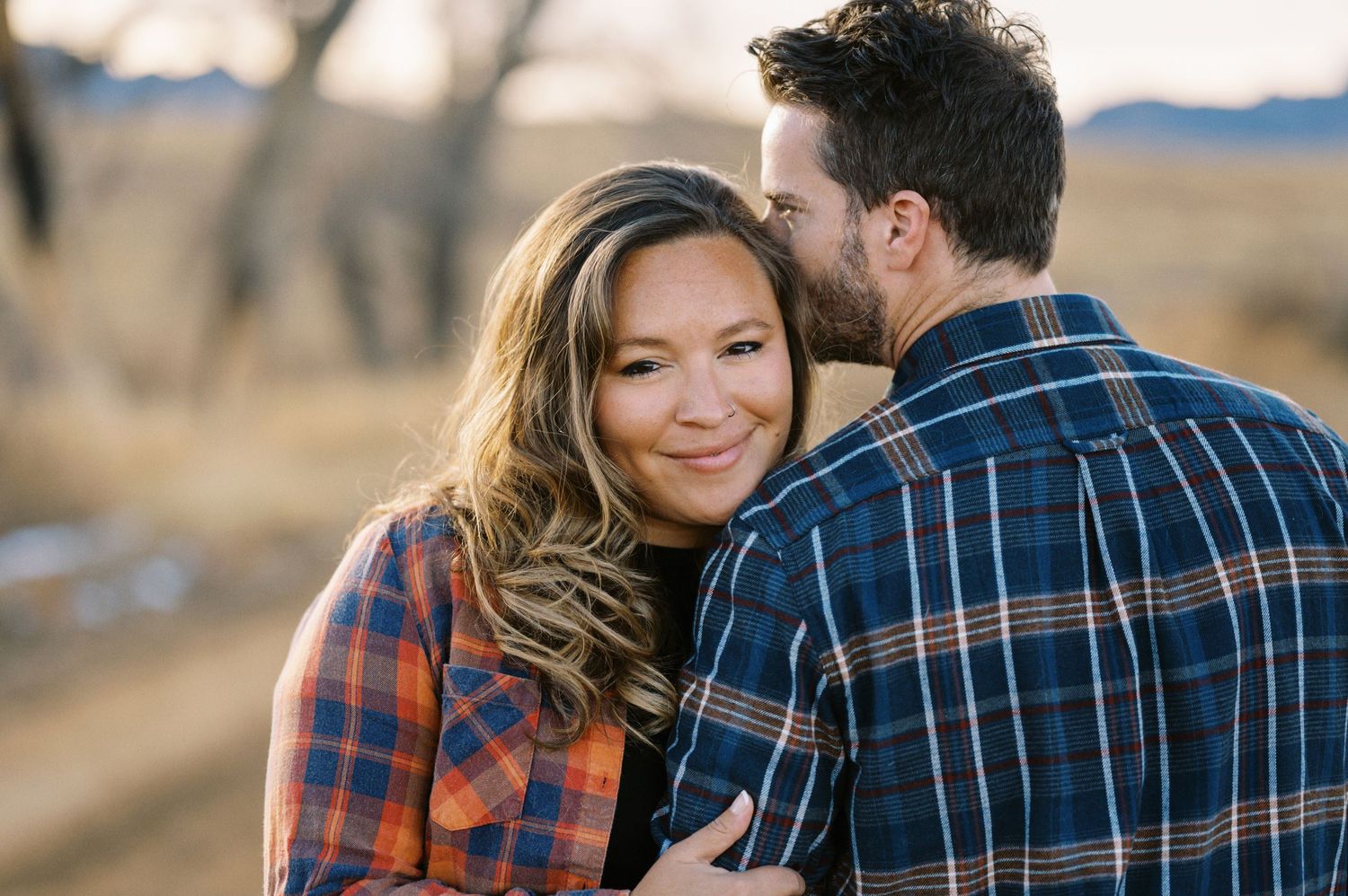A romantic couple wearing plaid shirts embraces on a rural road during golden hour.