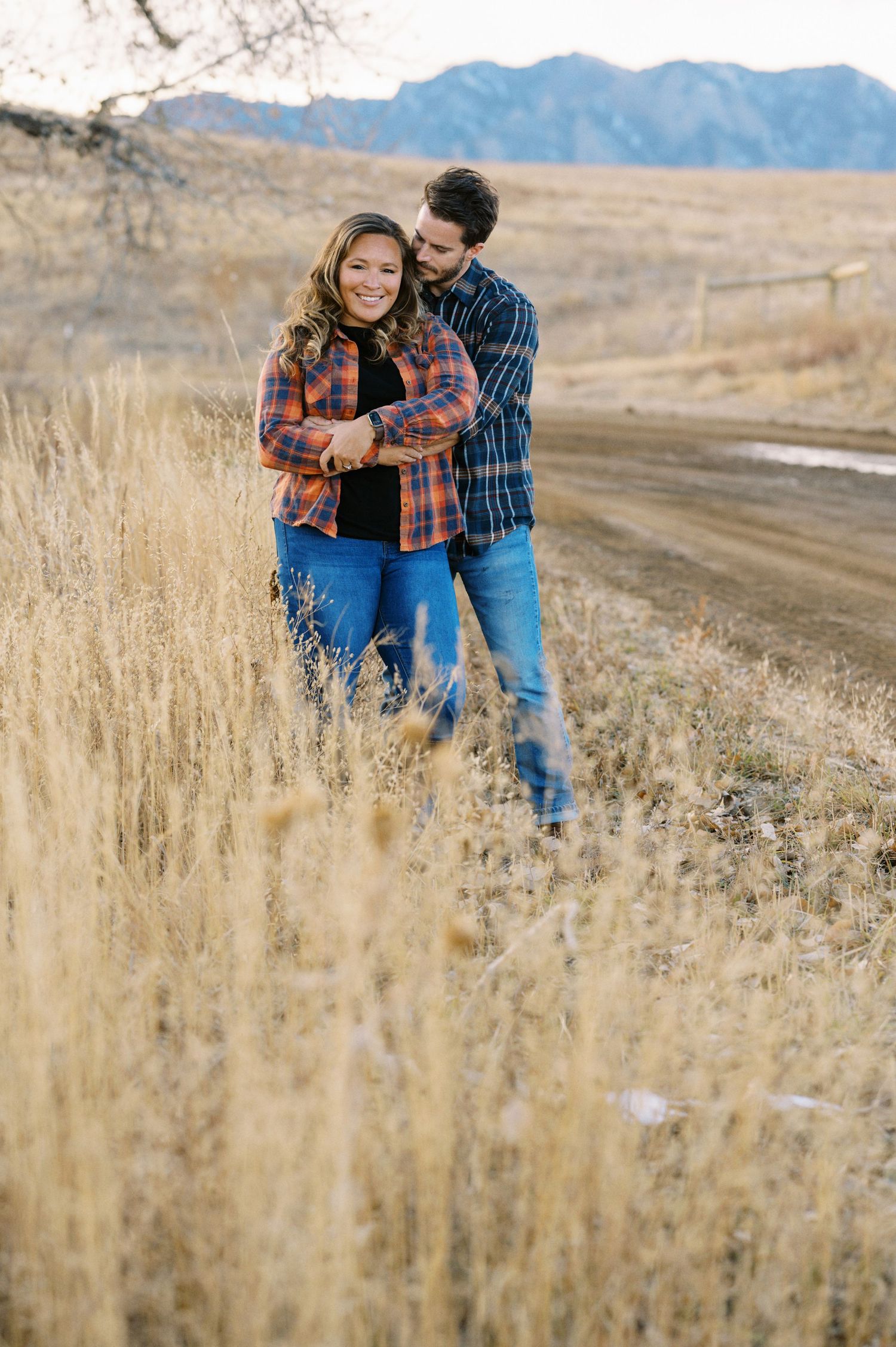A couple embraces in tall grass beside a dirt road with mountains in the distance.