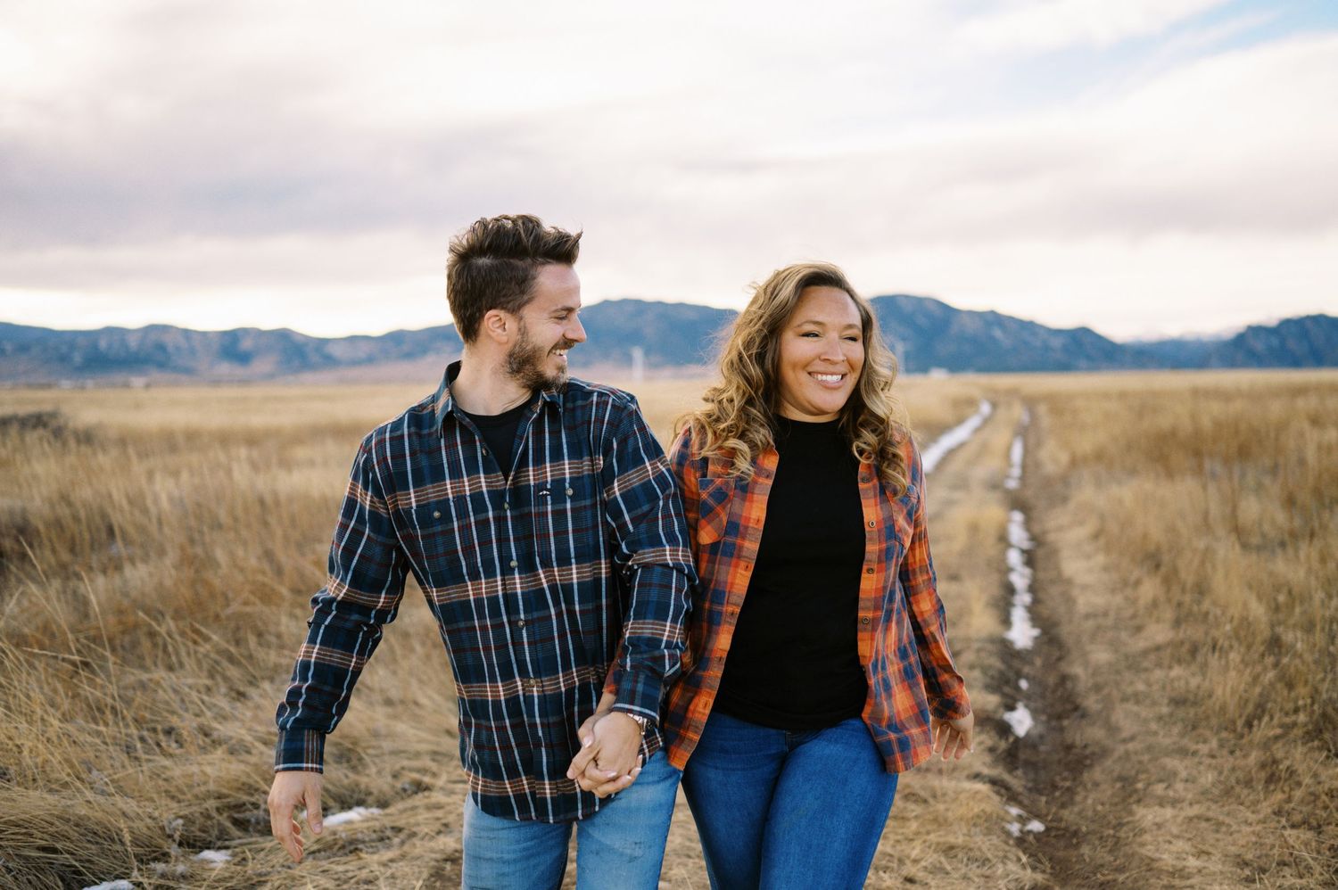 A couple walks hand in hand on a path through golden prairie grass with mountains in the distance.