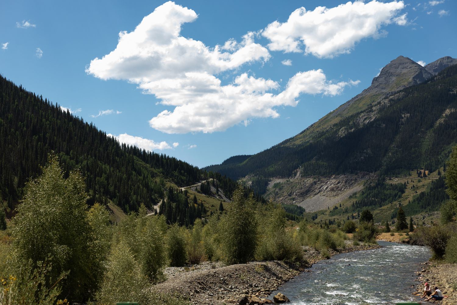 Scenic mountain landscape with a rushing river and forest under bright blue skies.