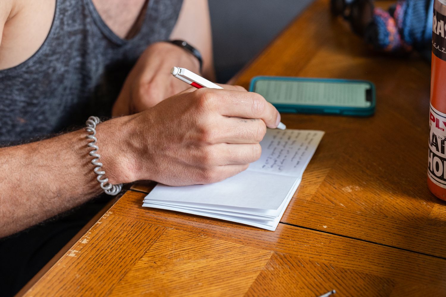 Close-up of hands writing in a notebook on a wooden table surface.