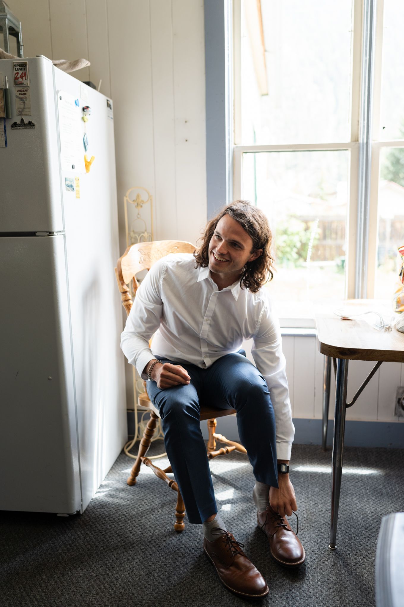 Groom getting ready with wooden chair and refrigerator near a bright window.