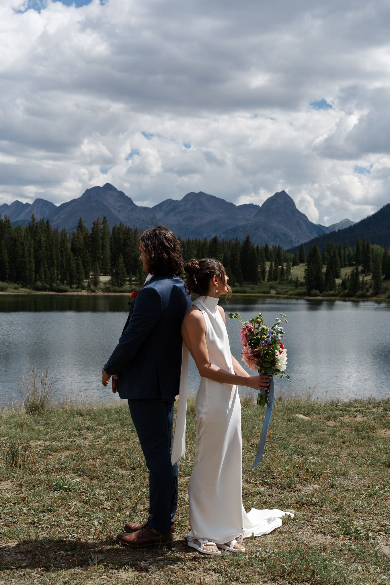 A couple in wedding attire stands by a mountain lake surrounded by evergreen trees and dramatic clouds.