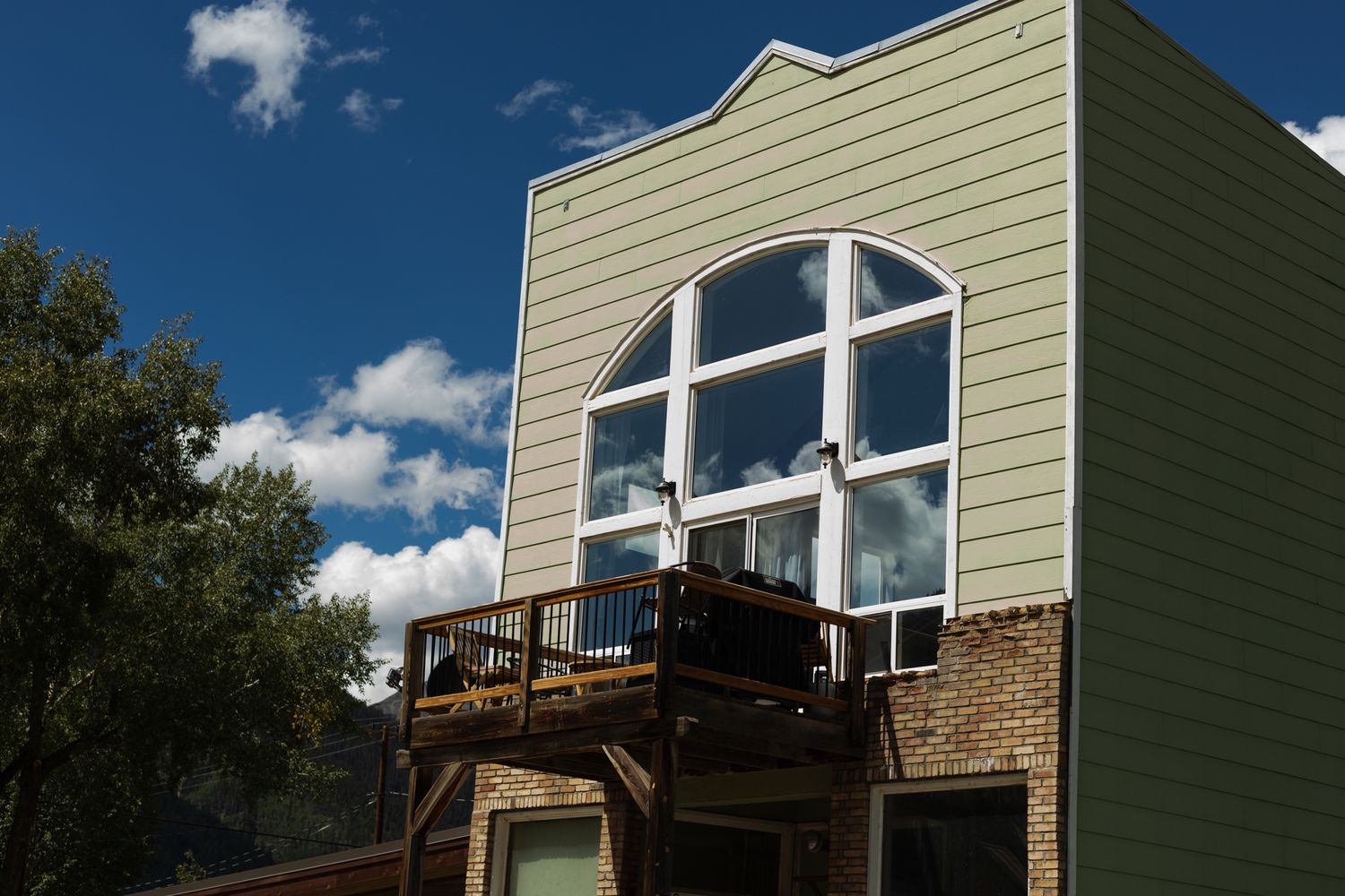Modern sage green house with large windows and a wooden deck against blue sky.