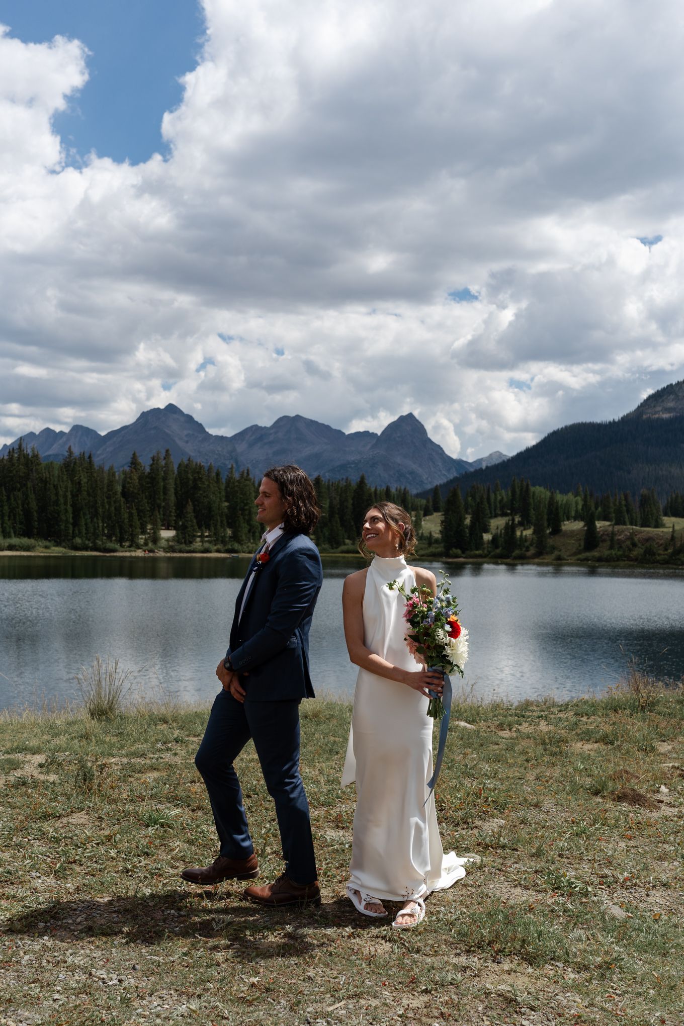 Wedding couple poses back-to-back by serene mountain lake with dramatic cloudy sky backdrop.