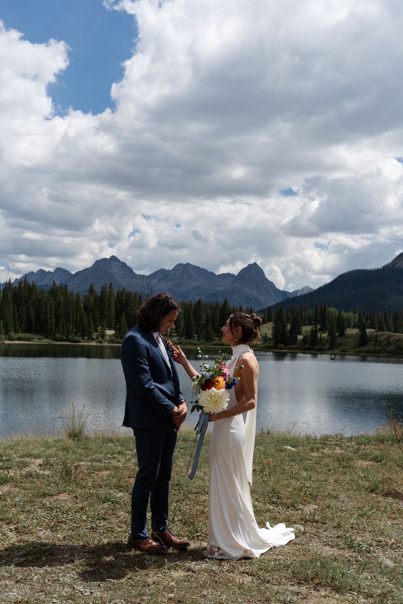 Intimate wedding moment captured lakeside with mountain peaks and forest in background.