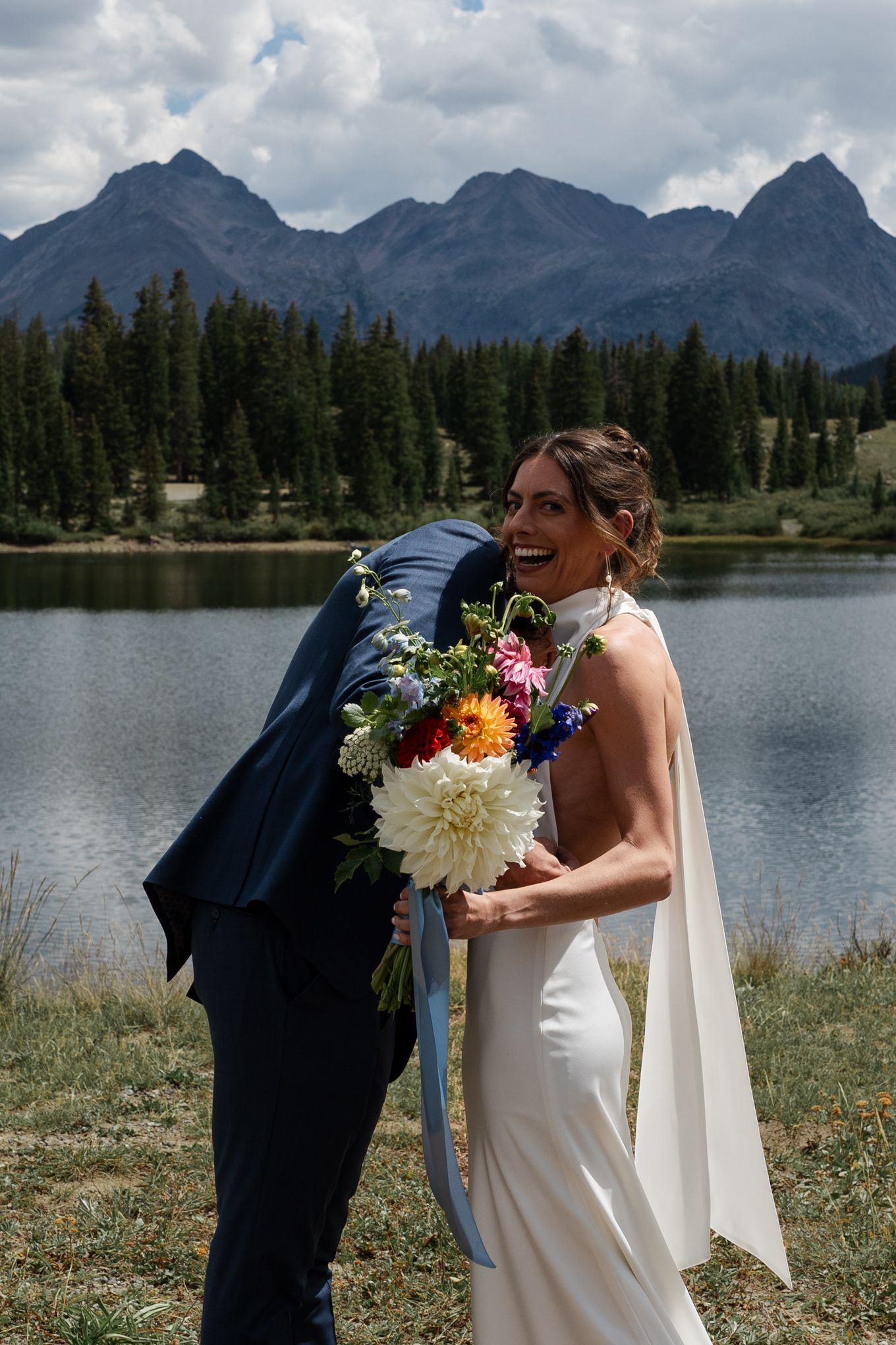 Joyful wedding couple embraces near a mountain lake with colorful bouquet and white dress.