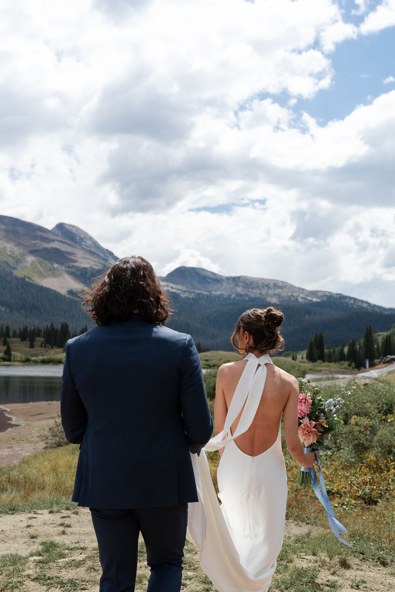 Couple in wedding attire views mountain landscape from behind near alpine lake.