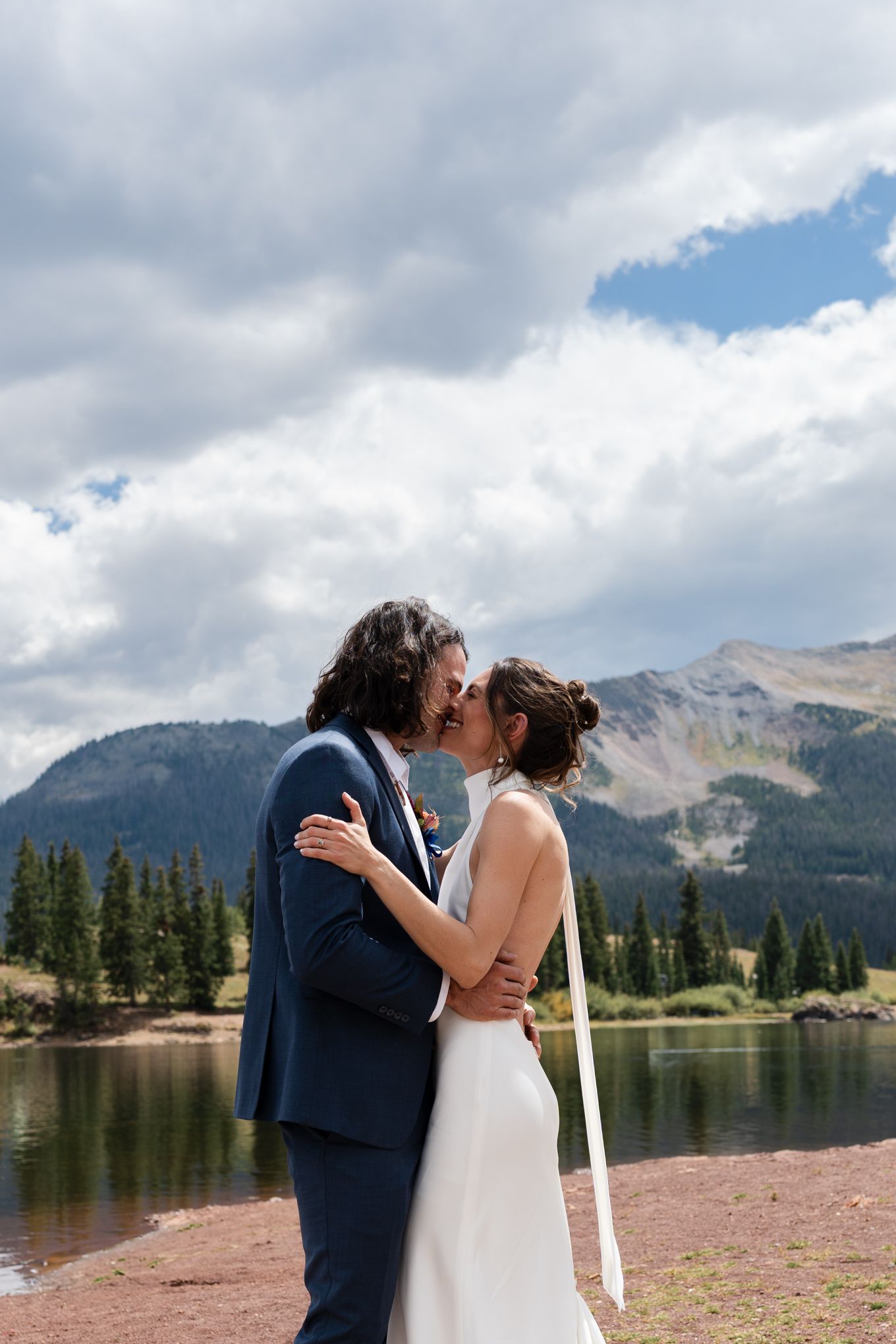 Couple shares romantic kiss against backdrop of mountain lake and cloudy sky.