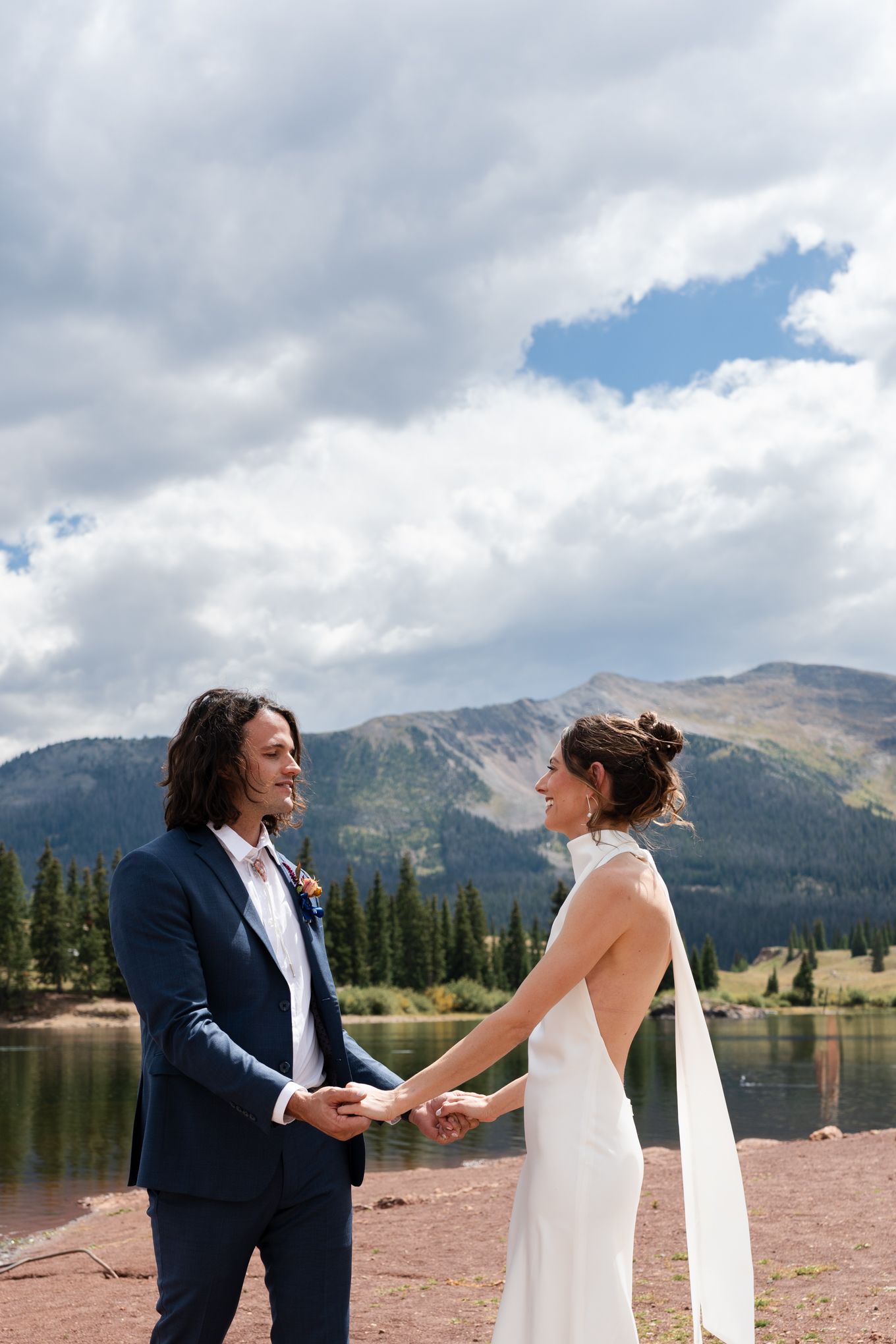 Wedding couple holds hands while standing by mountain lake with dramatic cloud formation.