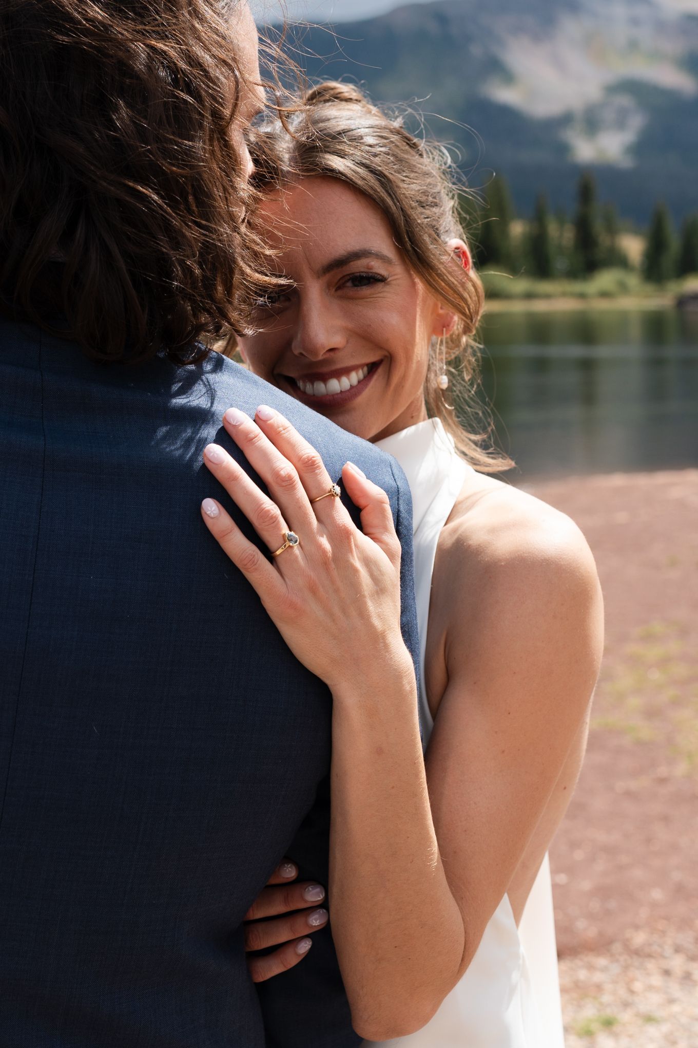 Close-up of embrace showing wedding ring detail and joyful expression by mountain lake.
