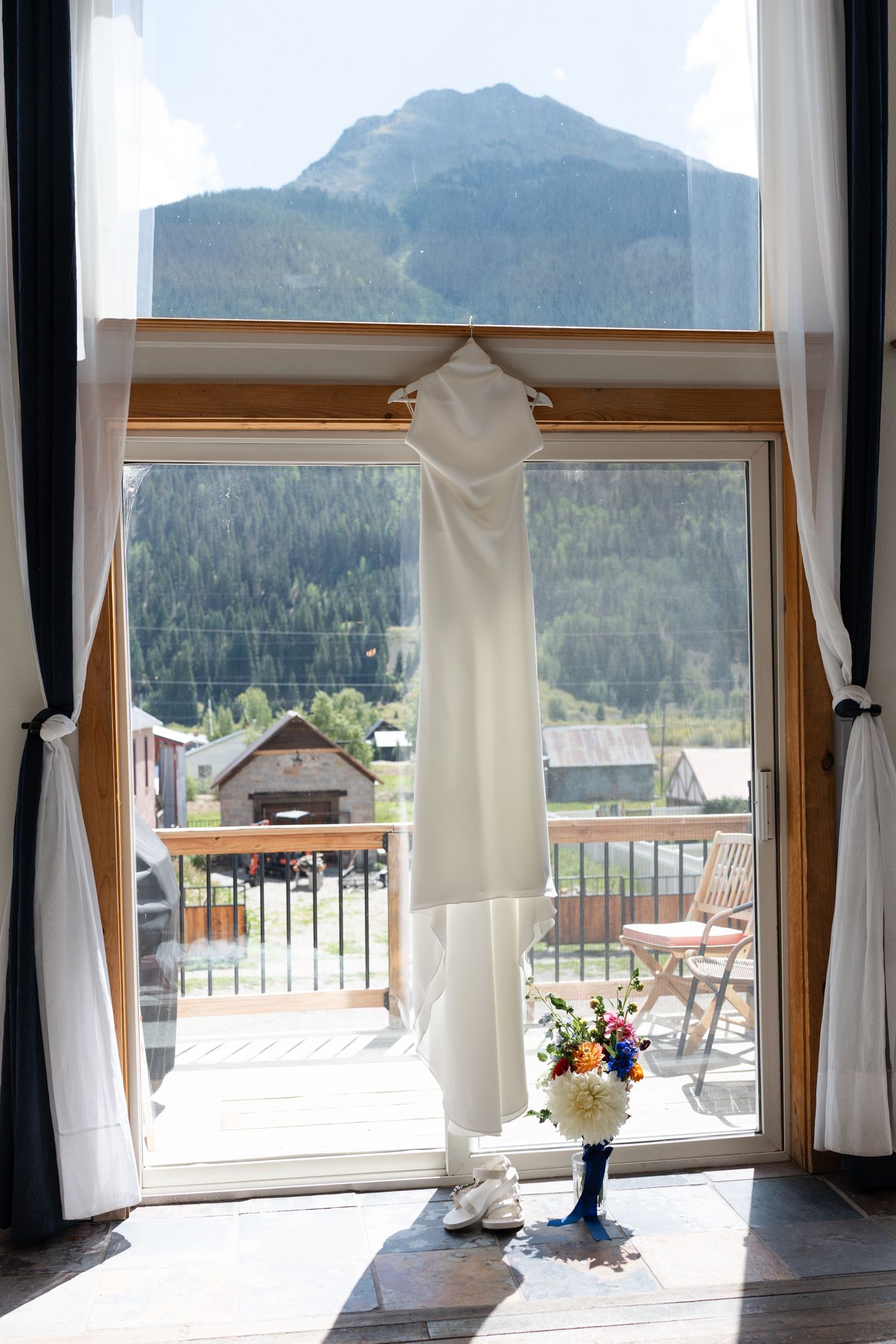 White wedding dress hanging in window frame with mountain views and balcony in background.