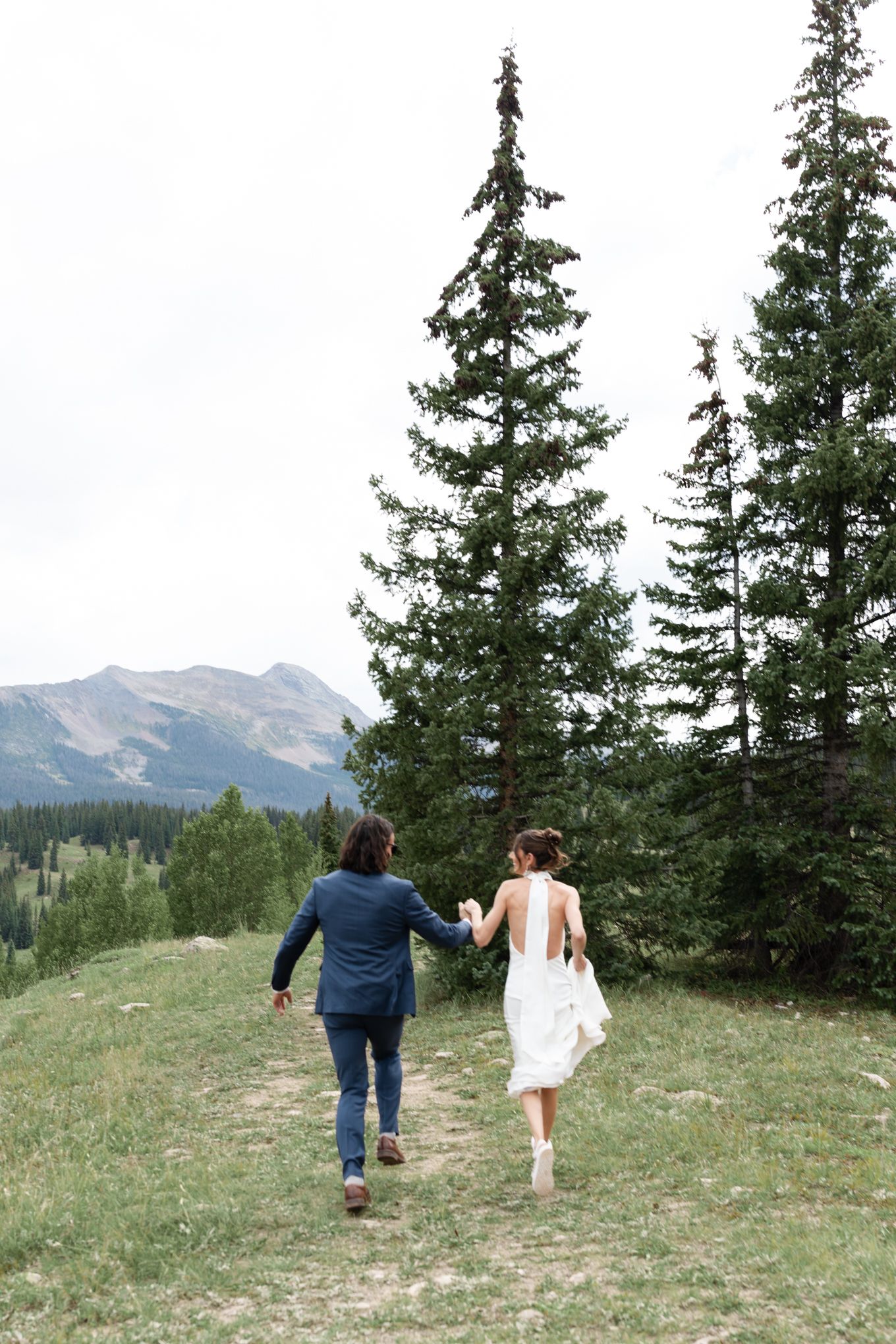Newlyweds running together on mountainside path with evergreen trees and peaks in background.