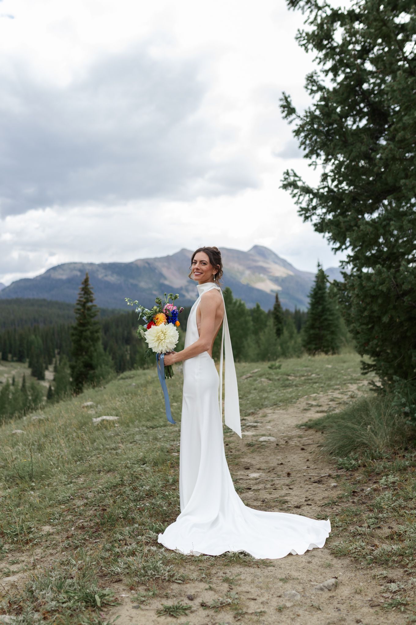Elegant bride in flowing dress holds bouquet while standing on mountain trail with scenic vista.