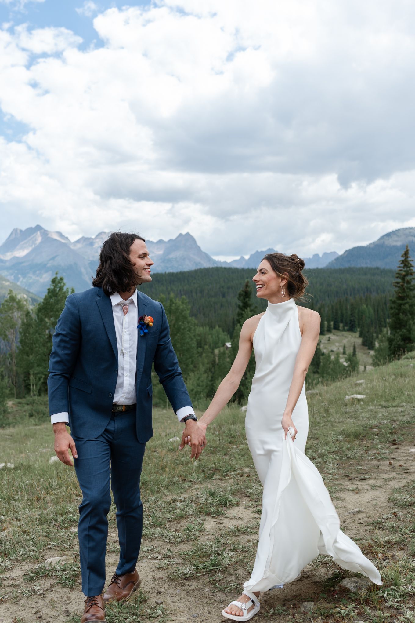 A couple in wedding attire walk hand in hand on a mountain trail with snow-capped peaks in the background.