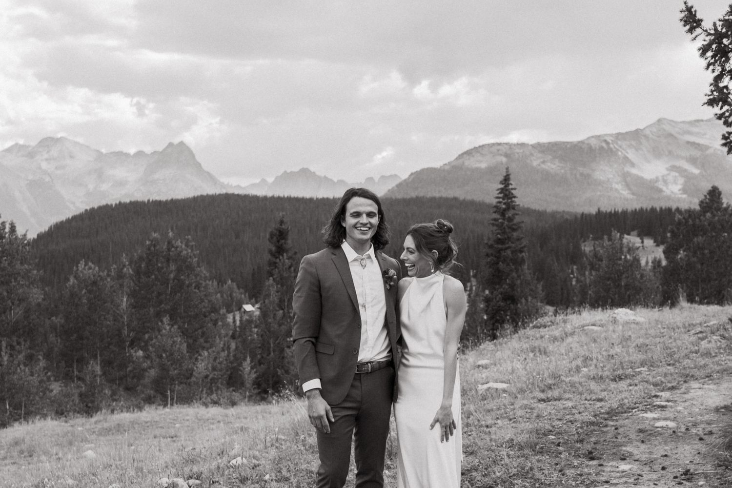 Black and white portrait of newlyweds against a dramatic mountain landscape with evergreen trees.