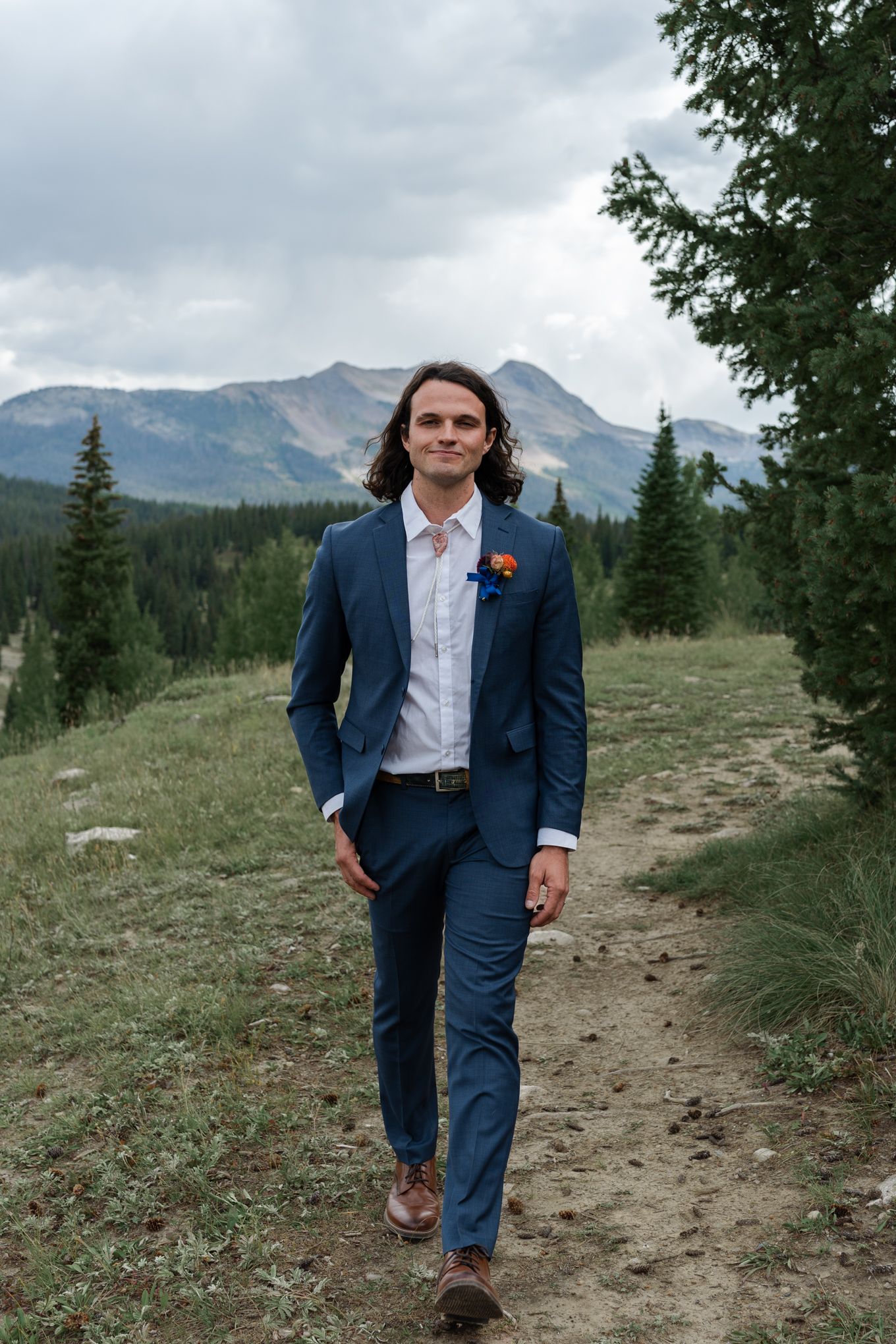 Groom in navy suit walks along mountain trail with evergreen forest and peaks in background.