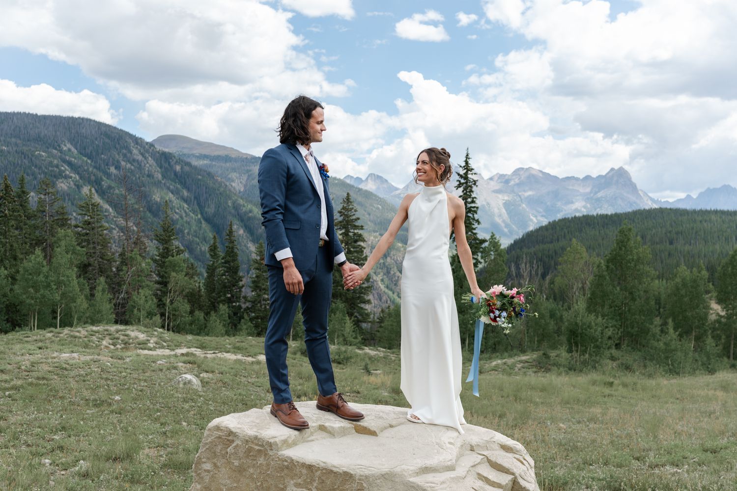 Newlyweds stand together on rocky outcrop with majestic mountain vista behind them.
