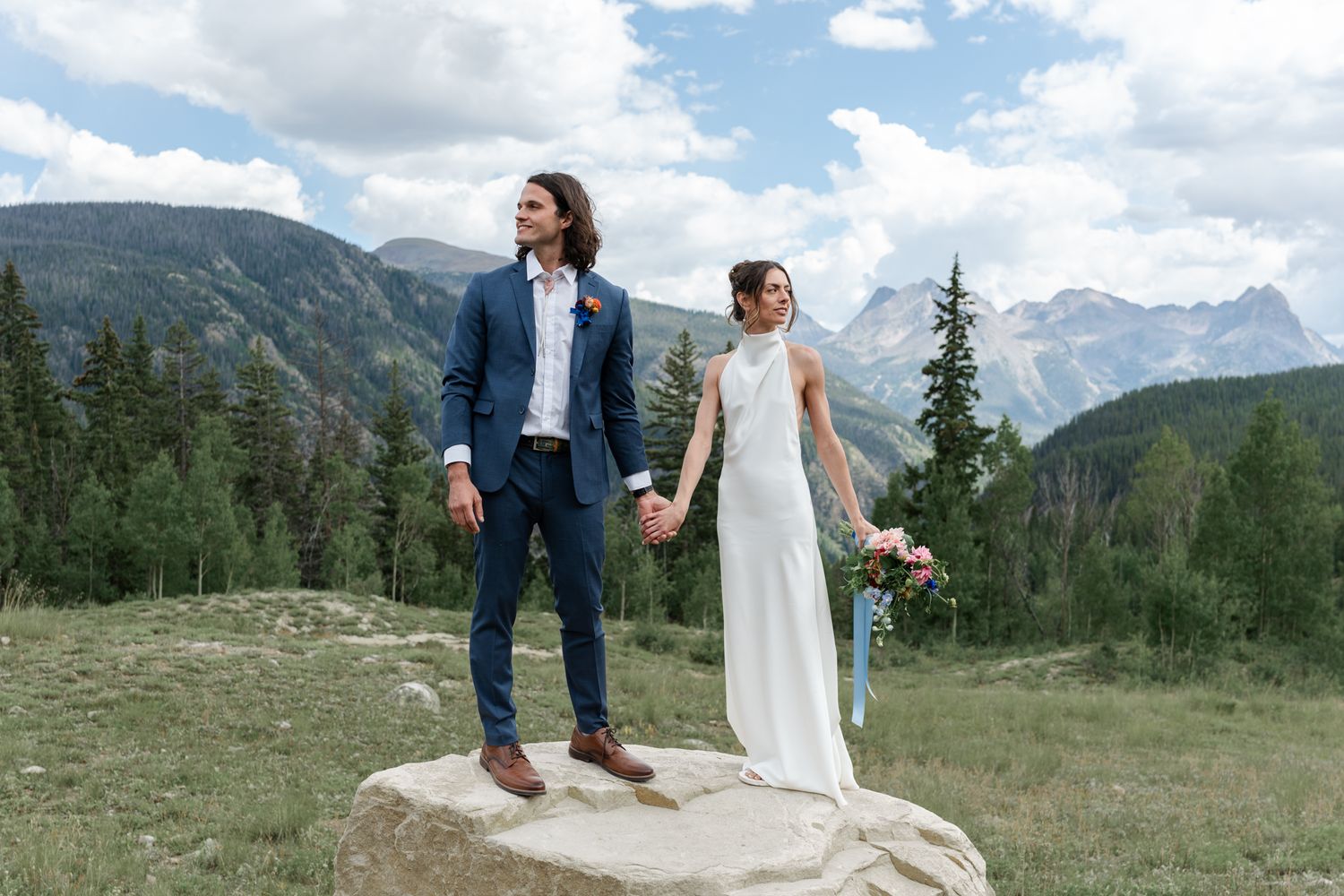 Wedding couple poses on large rock with dramatic mountain range and blue sky in background.