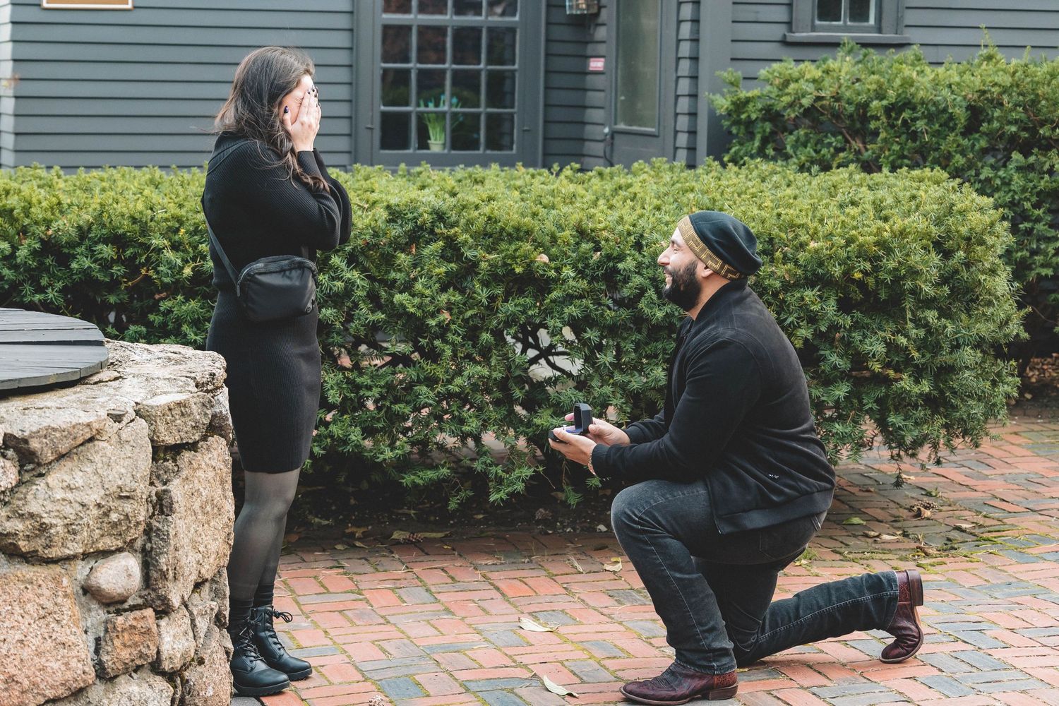 A person kneeling during a marriage proposal on brick pavement beside stone wall.