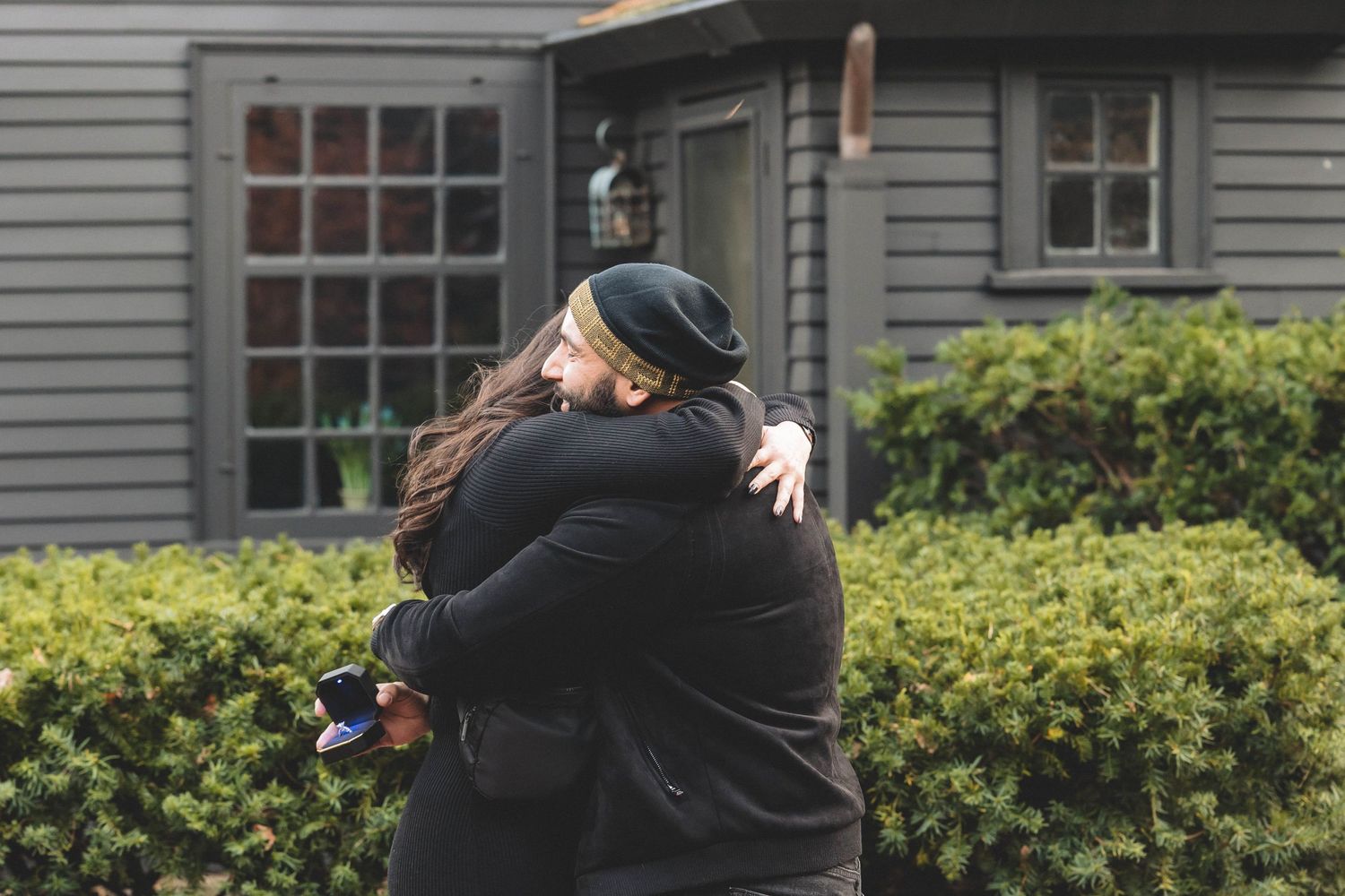 A couple embraces warmly in front of a grey house with bushes.